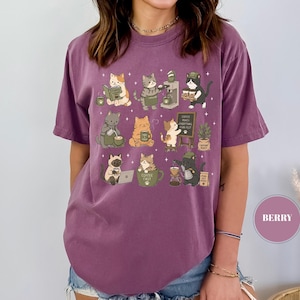 May include: Berry-colored t-shirt featuring a whimsical illustration of cats engaged in coffee-making activities. The design includes various cats, coffee machines, and the text "Coffee fuels everything, purr-fect". The shirt is made of a soft material.
