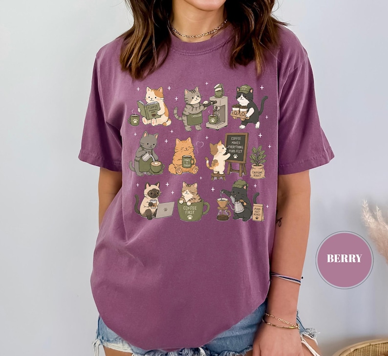 May include: Berry-colored t-shirt featuring a whimsical illustration of cats engaged in coffee-making activities. The design includes various cats, coffee machines, and the text "Coffee fuels everything, purr-fect". The shirt is made of a soft material.