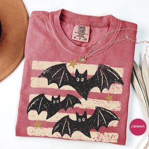 May include: A crimson short-sleeved t-shirt featuring a graphic print of three black bats flying over horizontal cream-colored distressed stripes and small gold stars. The shirt has a Comfort Colors brand label at the neck and a size small tag.