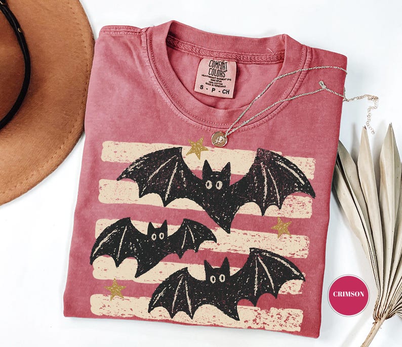 May include: A crimson short-sleeved t-shirt featuring a graphic print of three black bats flying over horizontal cream-colored distressed stripes and small gold stars. The shirt has a Comfort Colors brand label at the neck and a size small tag.