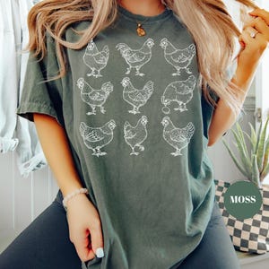May include: A dark green t-shirt featuring a white illustration of nine chickens in various poses. The shirt has a relaxed fit and a crew neck. The word "MOSS" is printed on a green circle.