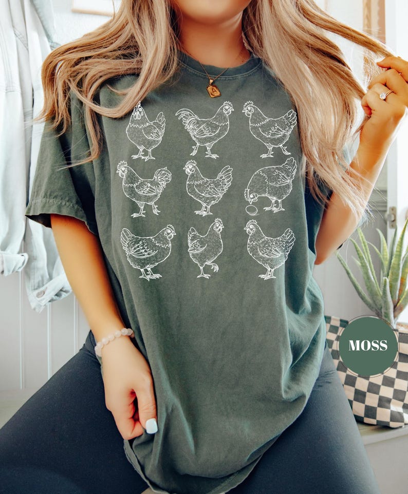 May include: A dark green t-shirt featuring a white illustration of nine chickens in various poses. The shirt has a relaxed fit and a crew neck. The word "MOSS" is printed on a green circle.