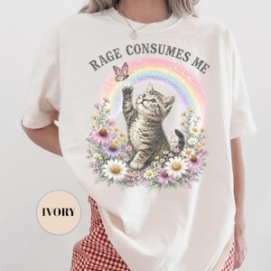 May include: Ivory-colored t-shirt featuring a graphic of a kitten reaching for a butterfly above a rainbow and flowers. The text "RAGE CONSUMES ME" is arched above the design. The shirt has short sleeves.