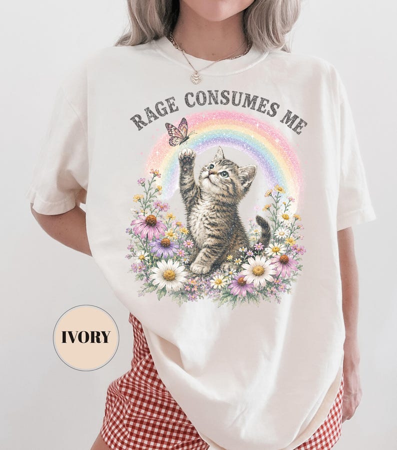 May include: Ivory-colored t-shirt featuring a graphic of a kitten reaching for a butterfly above a rainbow and flowers. The text "RAGE CONSUMES ME" is arched above the design. The shirt has short sleeves.