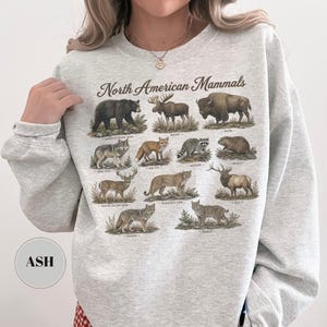 May include: A light gray crewneck sweatshirt featuring a graphic print titled North American Mammals. The design displays detailed illustrations of various animals including a black bear, moose, bison, gray wolf, red fox, raccoon, beaver, deer, mountain lion, elk, coyote, and bobcat, each labeled with its common name.