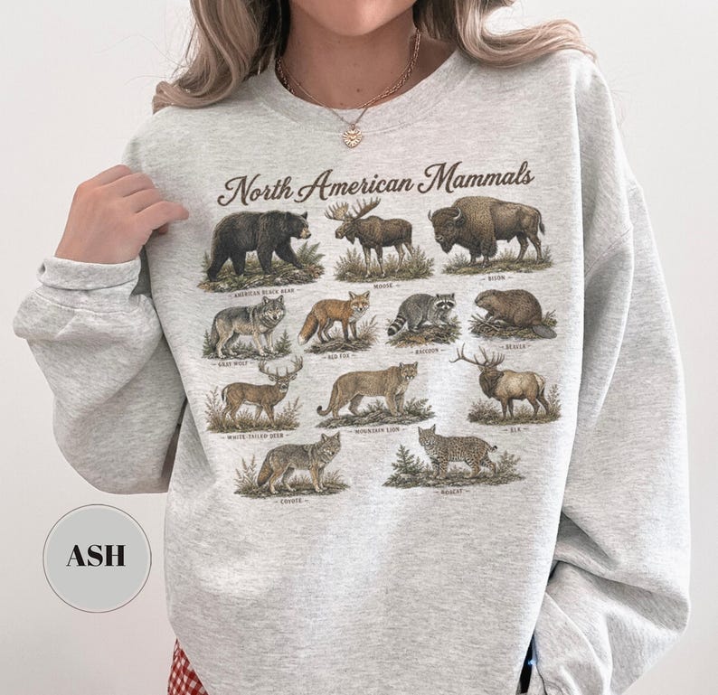 May include: A light gray crewneck sweatshirt featuring a graphic print titled North American Mammals. The design displays detailed illustrations of various animals including a black bear, moose, bison, gray wolf, red fox, raccoon, beaver, deer, mountain lion, elk, coyote, and bobcat, each labeled with its common name.