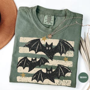 May include: A moss green short-sleeve t-shirt featuring a graphic print of three black bats flying over horizontal cream-colored distressed stripes and small gold stars. The inner neck label reads Comfort Colors, S-P-CH, 100% cotton.