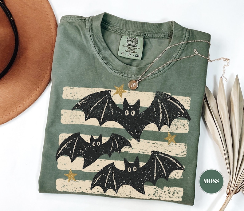 May include: A moss green short-sleeve t-shirt featuring a graphic print of three black bats flying over horizontal cream-colored distressed stripes and small gold stars. The inner neck label reads Comfort Colors, S-P-CH, 100% cotton.
