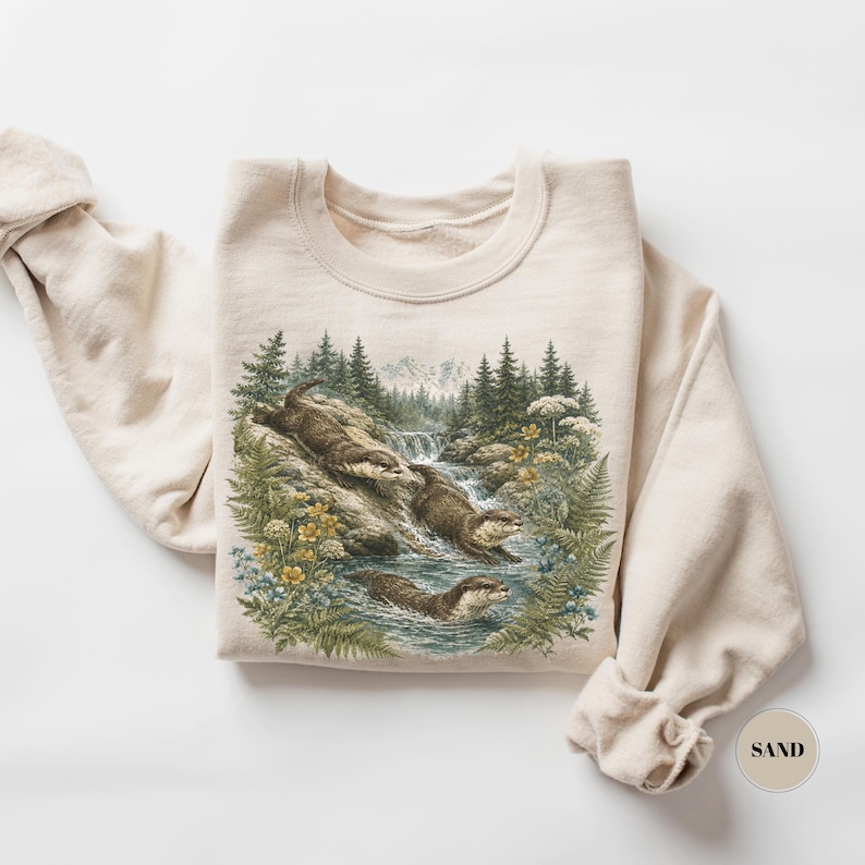 Puede incluir: Una sudadera de cuello redondo color arena con una ilustraci&oacute;n gr&aacute;fica detallada de tres nutrias en un entorno fluvial natural con pinos, helechos, flores silvestres amarillas y una cascada. El texto SAND aparece impreso en un peque&ntilde;o c&iacute;rculo.
