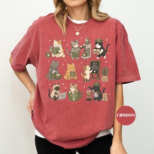 May include: Crimson-colored t-shirt featuring a whimsical design of various cats engaged in coffee-related activities. The cats are depicted brewing coffee, reading books, and enjoying their coffee. The phrase "Coffee Makes Everything Purr-fect" is on a small chalkboard.