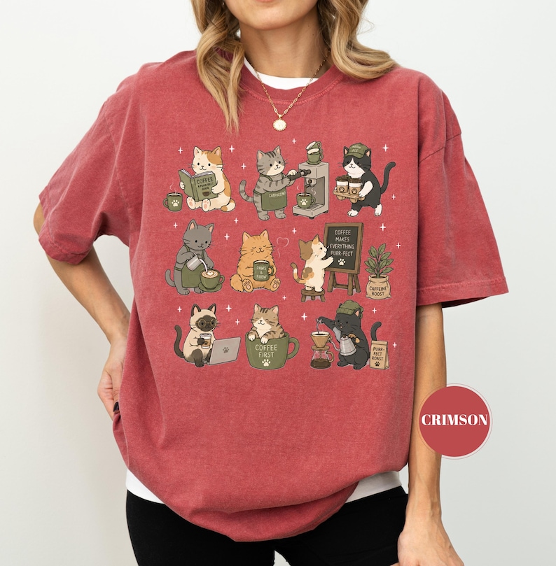 May include: Crimson-colored t-shirt featuring a whimsical design of various cats engaged in coffee-related activities. The cats are depicted brewing coffee, reading books, and enjoying their coffee. The phrase "Coffee Makes Everything Purr-fect" is on a small chalkboard.