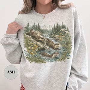 Puede incluir: Una sudadera de cuello redondo gris claro con un estampado gr&aacute;fico detallado de tres nutrias en un arroyo forestal. La ilustraci&oacute;n muestra a las nutrias nadando y trepando por rocas rodeadas de pinos, helechos y flores silvestres amarillas. El texto ASH es visible en un peque&ntilde;o c&iacute;rculo en la parte inferior izquierda.