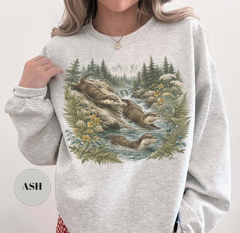 Puede incluir: Una sudadera de cuello redondo gris claro con un estampado gr&aacute;fico detallado de tres nutrias en un arroyo forestal. La ilustraci&oacute;n muestra a las nutrias nadando y trepando por rocas rodeadas de pinos, helechos y flores silvestres amarillas. El texto ASH es visible en un peque&ntilde;o c&iacute;rculo en la parte inferior izquierda.