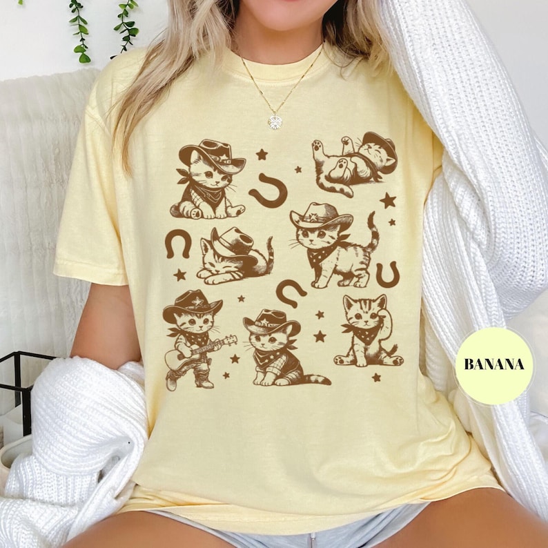 May include: Light yellow t-shirt featuring a repeating pattern of cartoon cowboy cats in various poses. The cats wear cowboy hats and bandanas, with horseshoes and stars. The word "BANANA" is printed on a yellow circle.