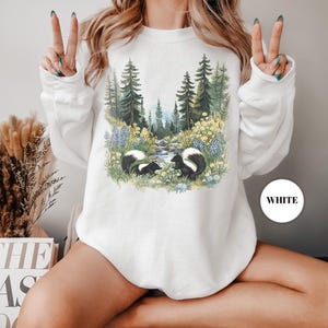 May include: White sweatshirt featuring a nature scene with two skunks, a stream, and a forest. The design includes blue and yellow flowers, and evergreen trees. The word "WHITE" is printed on a white circle.