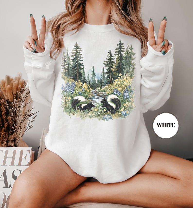 May include: White sweatshirt featuring a nature scene with two skunks, a stream, and a forest. The design includes blue and yellow flowers, and evergreen trees. The word "WHITE" is printed on a white circle.