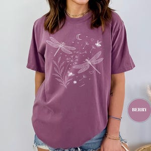 May include: Berry-colored t-shirt featuring a white dragonfly and floral design. The short-sleeved shirt has a crew neck and a relaxed fit. The graphic includes dragonflies, a crescent moon, bees, and hearts.