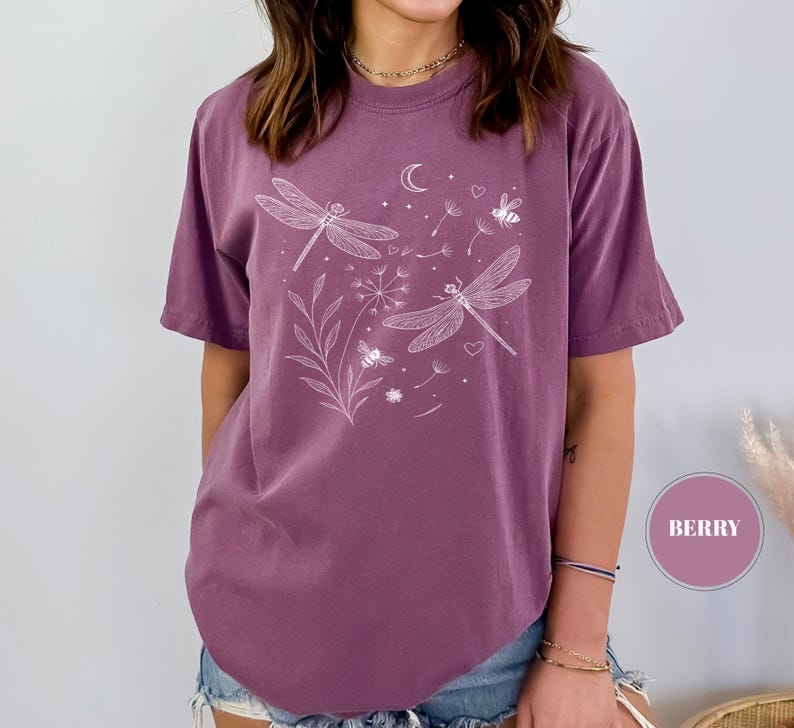 May include: Berry-colored t-shirt featuring a white dragonfly and floral design. The short-sleeved shirt has a crew neck and a relaxed fit. The graphic includes dragonflies, a crescent moon, bees, and hearts.