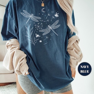 May include: Navy blue t-shirt with a white dragonfly and bee graphic. The design includes a crescent moon, dandelion seeds, and hearts. The shirt has a relaxed fit and the words "NAVY BLUE" are in a navy blue circle.