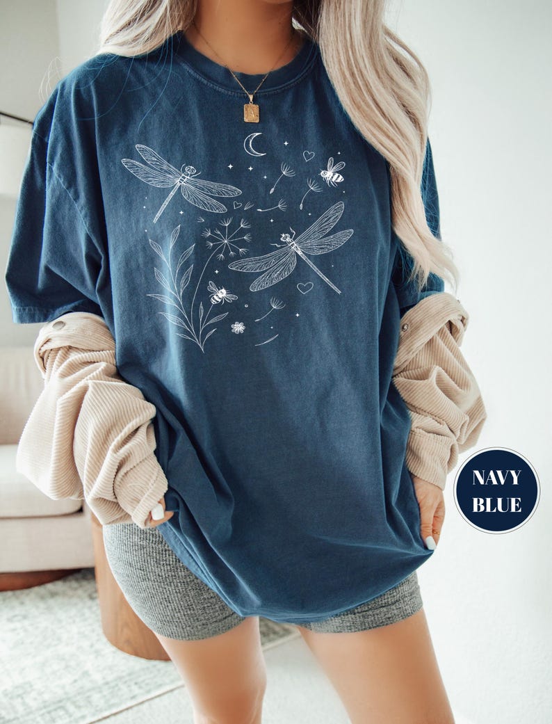 May include: Navy blue t-shirt with a white dragonfly and bee graphic. The design includes a crescent moon, dandelion seeds, and hearts. The shirt has a relaxed fit and the words "NAVY BLUE" are in a navy blue circle.