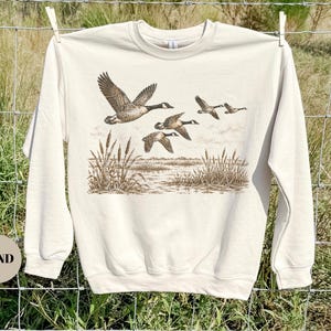 Peut inclure: Un sweat-shirt &agrave; col rond couleur sable arborant une impression graphique marron d&eacute;taill&eacute;e repr&eacute;sentant un vol d'oies au-dessus d'un paysage mar&eacute;cageux avec des massettes. Le pull &agrave; manches longues poss&egrave;de un col, des poignets et un ourlet c&ocirc;tel&eacute;s.