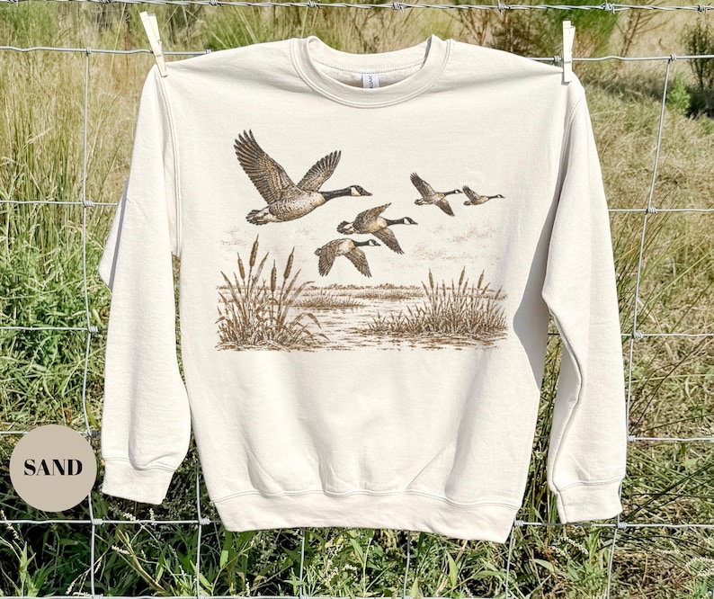 Peut inclure: Un sweat-shirt &agrave; col rond couleur sable arborant une impression graphique marron d&eacute;taill&eacute;e repr&eacute;sentant un vol d'oies au-dessus d'un paysage mar&eacute;cageux avec des massettes. Le pull &agrave; manches longues poss&egrave;de un col, des poignets et un ourlet c&ocirc;tel&eacute;s.
