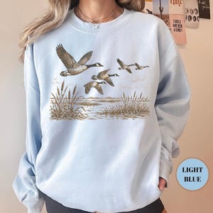 Peut inclure: Un sweat-shirt bleu clair &agrave; col rond orn&eacute; d'une illustration repr&eacute;sentant quatre oies volant au-dessus d'un paysage mar&eacute;cageux avec des roseaux. Le motif est imprim&eacute; dans des tons de brun et de beige sur le tissu bleu clair uni.