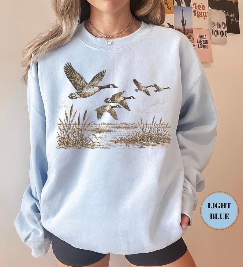 Peut inclure: Un sweat-shirt bleu clair &agrave; col rond orn&eacute; d'une illustration repr&eacute;sentant quatre oies volant au-dessus d'un paysage mar&eacute;cageux avec des roseaux. Le motif est imprim&eacute; dans des tons de brun et de beige sur le tissu bleu clair uni.