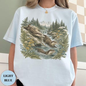 Puede incluir: Una camiseta azul claro con un estampado gr&aacute;fico detallado de tres nutrias marrones nadando en un arroyo. La escena incluye helechos verdes, flores silvestres amarillas y un fondo de bosque con monta&ntilde;as bajo un cielo despejado, estilo vintage.