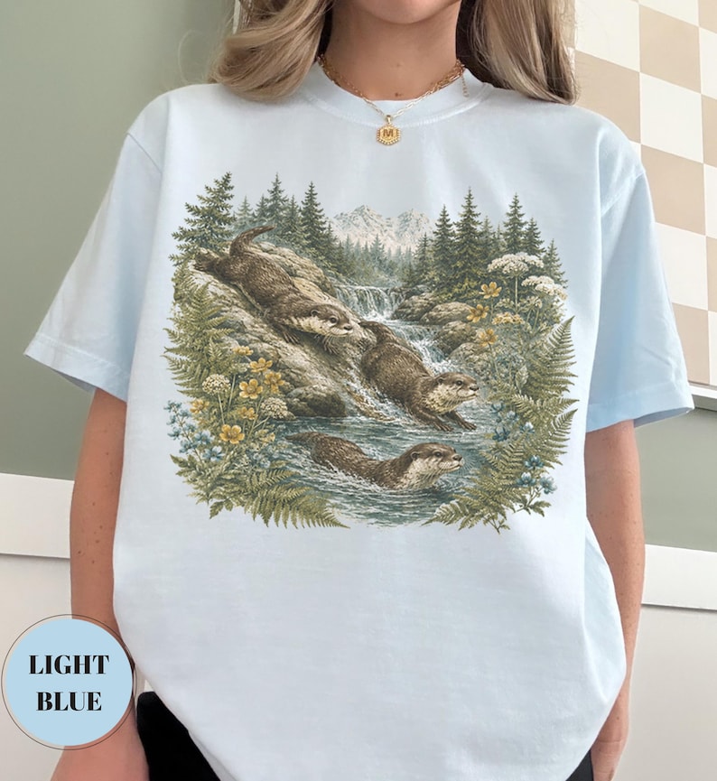 Puede incluir: Una camiseta azul claro con un estampado gr&aacute;fico detallado de tres nutrias marrones nadando en un arroyo. La escena incluye helechos verdes, flores silvestres amarillas y un fondo de bosque con monta&ntilde;as bajo un cielo despejado, estilo vintage.