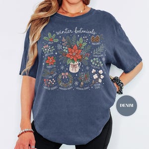 May include: A denim blue short-sleeve t-shirt featuring a graphic print titled winter botanicals. The design displays various holiday plants including holly, mistletoe, poinsettia, pinecones, cedar greens, eucalyptus, pine sprigs, winter berries, an evergreen wreath, a botanical bundle, and cotton stems.
