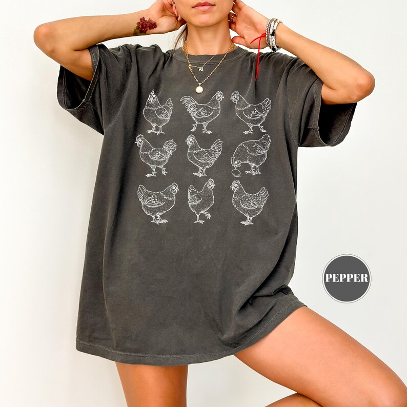 May include: Dark gray t-shirt featuring a grid of nine white chicken illustrations. The shirt has a relaxed fit and short sleeves. The word "PEPPER" is printed on a small, circular tag on the lower right side of the shirt.