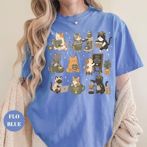 May include: A blue t-shirt featuring a whimsical design of various cats engaged in coffee-related activities. The cats are depicted in different poses, some reading, brewing coffee, or enjoying a cup. The shirt has the text "Coffee First".
