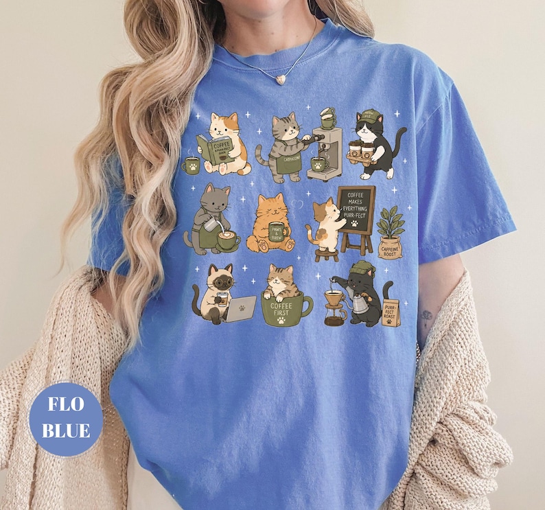 May include: A blue t-shirt featuring a whimsical design of various cats engaged in coffee-related activities. The cats are depicted in different poses, some reading, brewing coffee, or enjoying a cup. The shirt has the text "Coffee First".