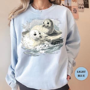 Pode incluir: Camisola azul clara com uma ilustra&ccedil;&atilde;o vintage de duas focas brancas com olhos escuros, sobre uma rocha no meio de ondas azuis e cinzentas. A camisola &eacute; de cor azul clara.