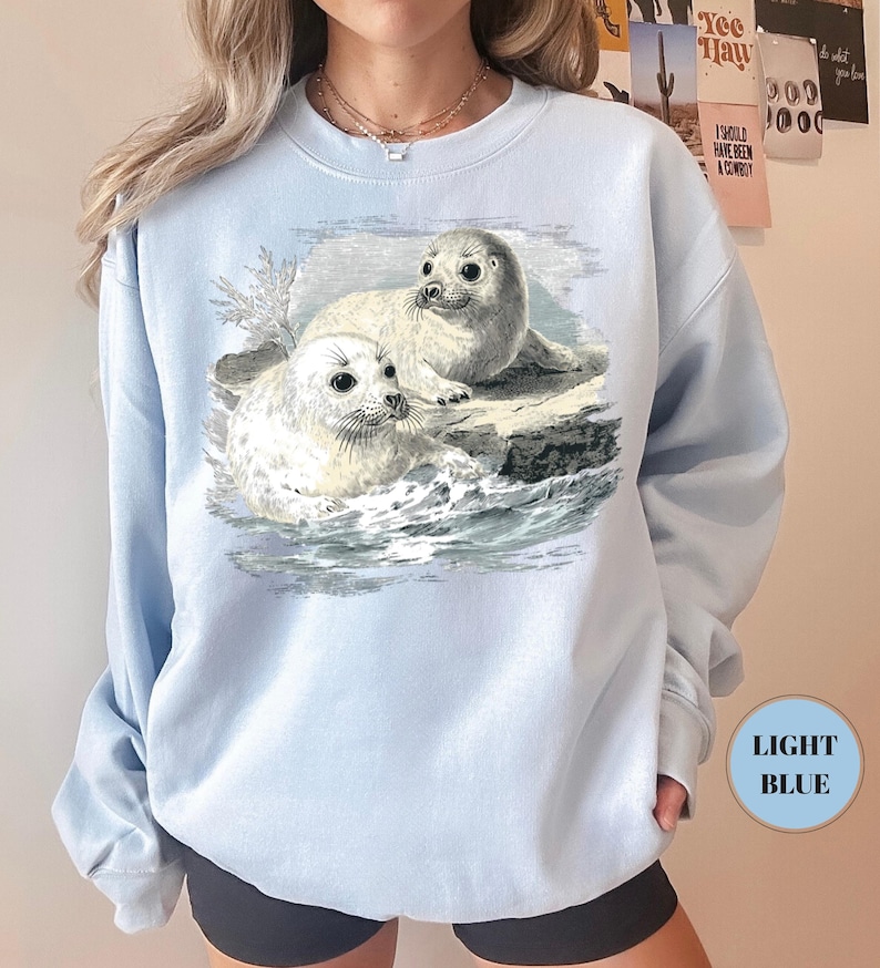 Pode incluir: Camisola azul clara com uma ilustra&ccedil;&atilde;o vintage de duas focas brancas com olhos escuros, sobre uma rocha no meio de ondas azuis e cinzentas. A camisola &eacute; de cor azul clara.