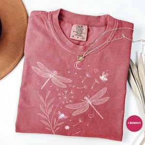May include: Crimson-colored Comfort Colors t-shirt with a white dragonfly and bee graphic. The shirt has a crew neck and short sleeves. A silver necklace with a moon pendant is draped on the shirt.