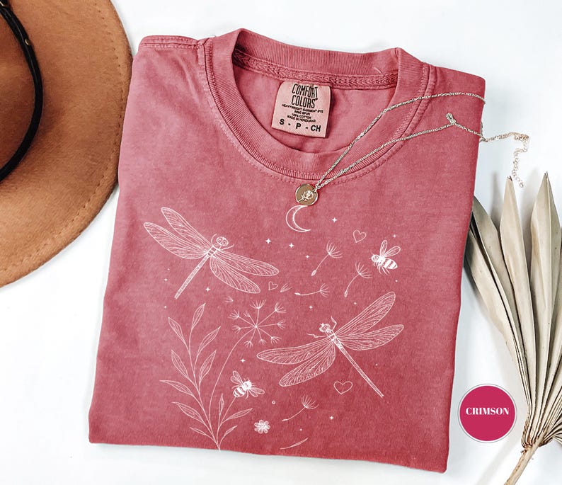 May include: Crimson-colored Comfort Colors t-shirt with a white dragonfly and bee graphic. The shirt has a crew neck and short sleeves. A silver necklace with a moon pendant is draped on the shirt.