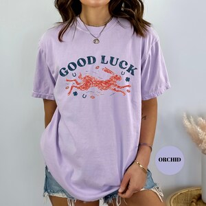 K&ouml;nnte beinhalten: Ein helllila T-Shirt mit den Worten "GOOD LUCK" in Dunkelblau &uuml;ber einer laufenden orangefarbenen Kaninchengrafik. Das Shirt zeigt auch orangefarbene Hufeisen und Kleebl&auml;tter. Das Wort "ORCHID" befindet sich in einem lila Kreis.