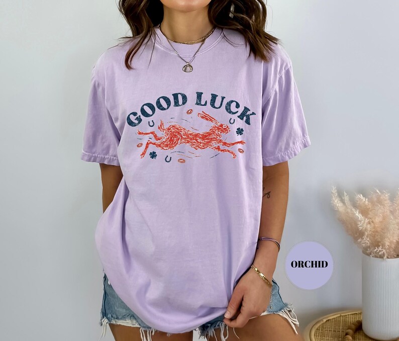 K&ouml;nnte beinhalten: Ein helllila T-Shirt mit den Worten "GOOD LUCK" in Dunkelblau &uuml;ber einer laufenden orangefarbenen Kaninchengrafik. Das Shirt zeigt auch orangefarbene Hufeisen und Kleebl&auml;tter. Das Wort "ORCHID" befindet sich in einem lila Kreis.