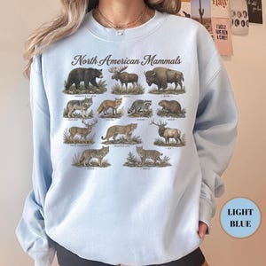 May include: A light blue crewneck sweatshirt featuring a graphic print titled North American Mammals. The design displays illustrations of a black bear, moose, bison, gray wolf, red fox, raccoon, beaver, white-tailed deer, mountain lion, elk, coyote, and bobcat.