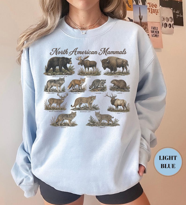 May include: A light blue crewneck sweatshirt featuring a graphic print titled North American Mammals. The design displays illustrations of a black bear, moose, bison, gray wolf, red fox, raccoon, beaver, white-tailed deer, mountain lion, elk, coyote, and bobcat.