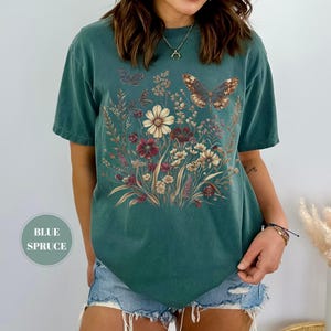 May include: A blue spruce colored short-sleeve t-shirt featuring a central graphic print of a wildflower bouquet in shades of cream, red, and brown, accented by two butterflies. The shirt has a crew neckline and a relaxed, boxy fit.