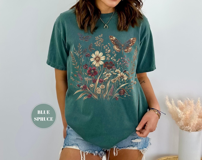 May include: A blue spruce colored short-sleeve t-shirt featuring a central graphic print of a wildflower bouquet in shades of cream, red, and brown, accented by two butterflies. The shirt has a crew neckline and a relaxed, boxy fit.