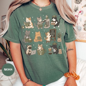 May include: Moss green t-shirt featuring a whimsical illustration of various cats engaged in coffee-related activities. The cats are depicted brewing coffee, reading, and enjoying coffee. The text "Coffee Makes Everything Purr-fect" is also visible.