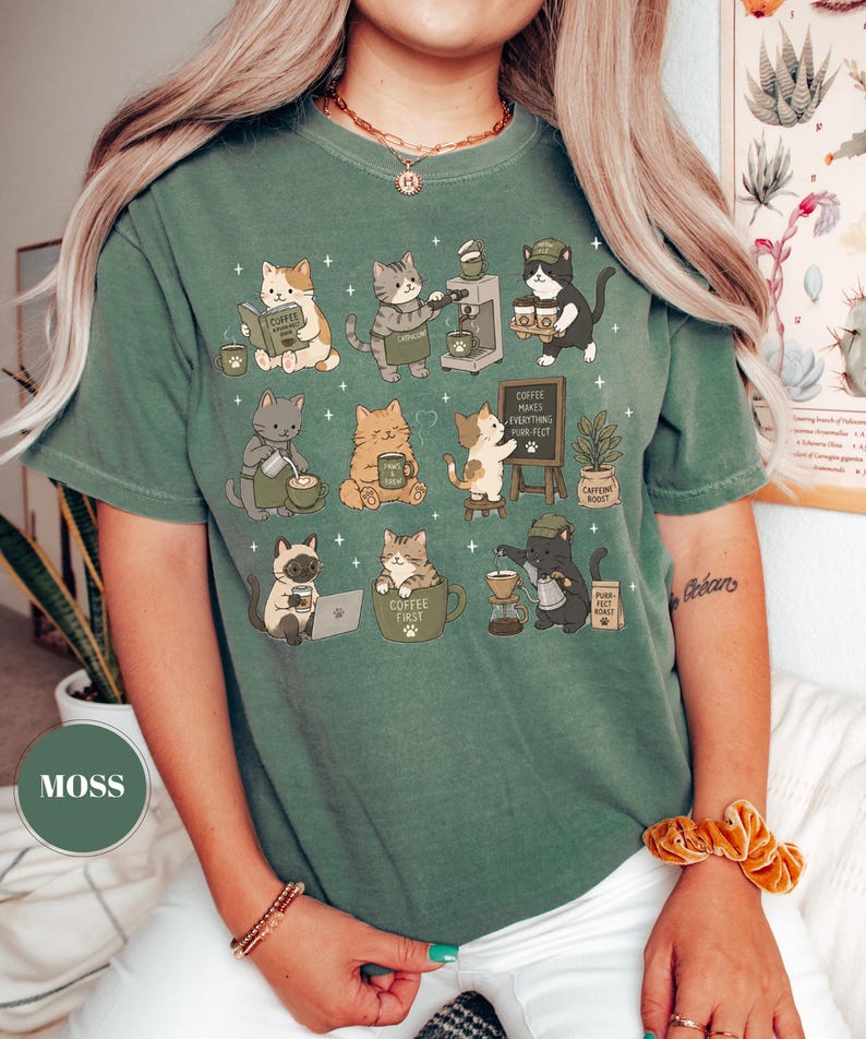 May include: Moss green t-shirt featuring a whimsical illustration of various cats engaged in coffee-related activities. The cats are depicted brewing coffee, reading, and enjoying coffee. The text "Coffee Makes Everything Purr-fect" is also visible.
