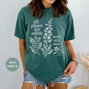 以下が含まれることがあります： ブルー・スプルース色のTシャツで、フォックスグローブの植物とベリーの白いボタニカルイラストが描かれています。前面には「all plants are edible some only once」というテキストがプリントされています。半袖でクルーネックのデザインです。