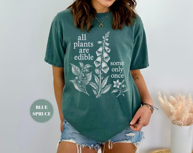 以下が含まれることがあります： ブルー・スプルース色のTシャツで、フォックスグローブの植物とベリーの白いボタニカルイラストが描かれています。前面には「all plants are edible some only once」というテキストがプリントされています。半袖でクルーネックのデザインです。