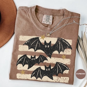 May include: A brown short-sleeve t-shirt featuring a graphic of three black bats flying over horizontal distressed cream-colored stripes and small gold stars. The shirt has a crew neckline and a Comfort Colors brand label on the inside collar.