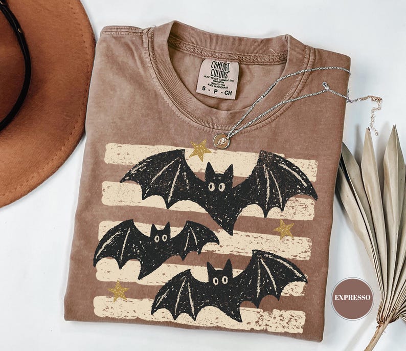 May include: A brown short-sleeve t-shirt featuring a graphic of three black bats flying over horizontal distressed cream-colored stripes and small gold stars. The shirt has a crew neckline and a Comfort Colors brand label on the inside collar.